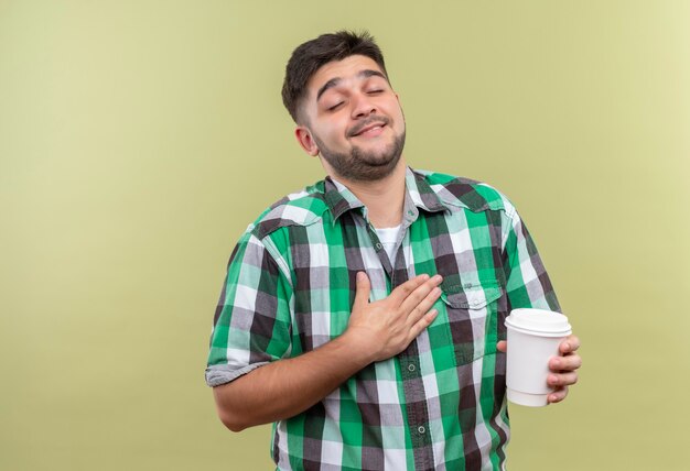 Jeune beau mec portant une chemise à carreaux pointant vers lui-même tenant une tasse de café en plastique debout sur un mur kaki