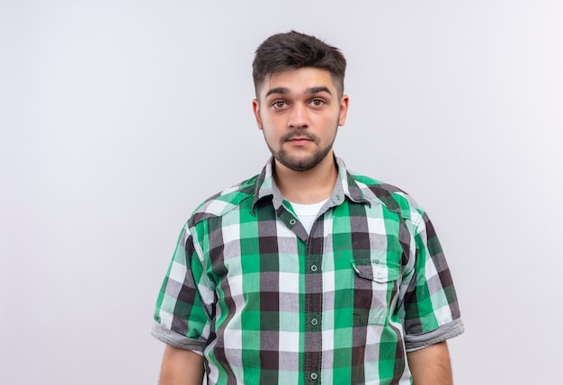 Jeune beau mec portant une chemise à carreaux à la recherche debout sans émotion sur un mur blanc