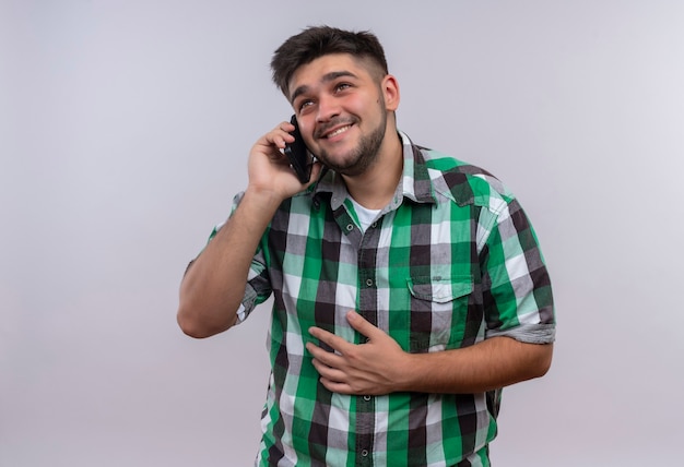 Jeune beau mec portant une chemise à carreaux en riant et en parlant au téléphone debout sur un mur blanc