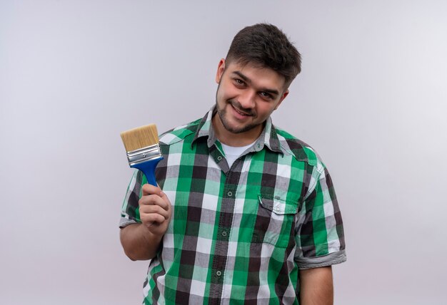 Jeune beau mec portant une chemise à carreaux souriant tenant un pinceau debout sur un mur blanc