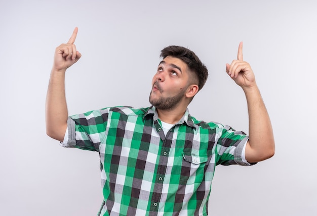 Jeune beau mec portant chemise à carreaux surpris pointant vers le haut avec l'index debout sur un mur blanc