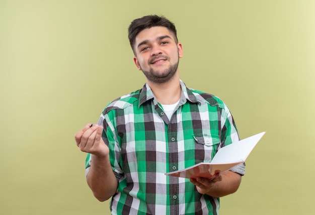 Jeune beau mec portant une chemise à carreaux tenant un stylo et un cahier debout sur un mur kaki