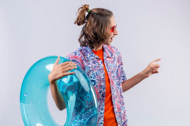 Jeune beau mec portant des lunettes de soleil rouges tenant un anneau gonflable à côté en pointant vers le côté avec le doigt debout