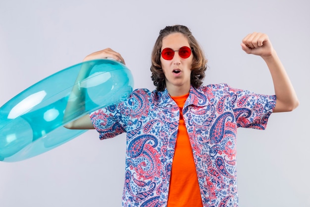 Jeune beau mec portant des lunettes de soleil rouges tenant un anneau gonflable levant le poing après une victoire sortie et heureux debout sur fond blanc