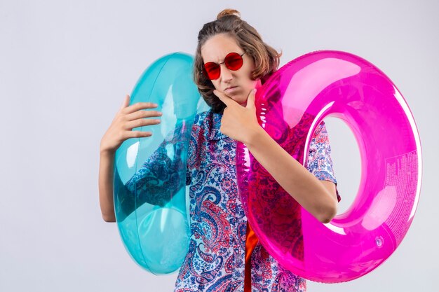 Jeune beau mec portant des lunettes de soleil rouges tenant des anneaux gonflables colorés regardant la caméra avec une expression suspecte pensive avec la main sur le menton pensant debout sur fond blanc