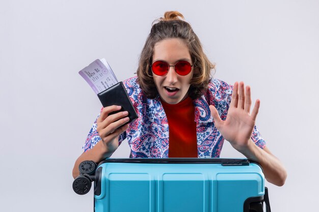 Jeune beau mec portant des lunettes de soleil rouges avec valise de voyage tenant des billets d'avion à la confiance et heureux debout sur fond bleu