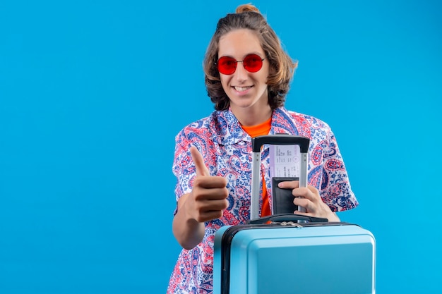 Jeune beau mec portant des lunettes de soleil rouges avec valise de voyage tenant des billets d'avion montrant les pouces vers le haut sourire heureux et positif joyeusement debout sur fond bleu