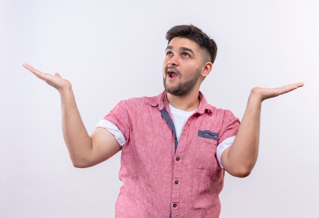 Jeune beau mec portant un polo rose en attente et levant les mains levées debout sur un mur blanc