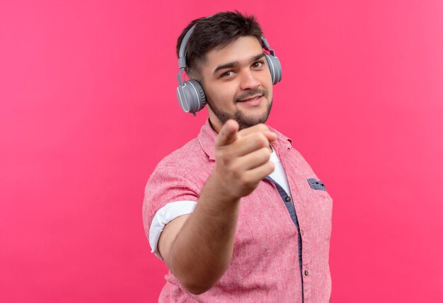 Jeune beau mec portant un polo rose et des écouteurs pointant joyeusement avec l'index debout sur le mur rose