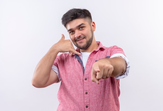 Jeune beau mec portant un polo rose faisant de manière ludique appelez-moi signe debout sur un mur blanc