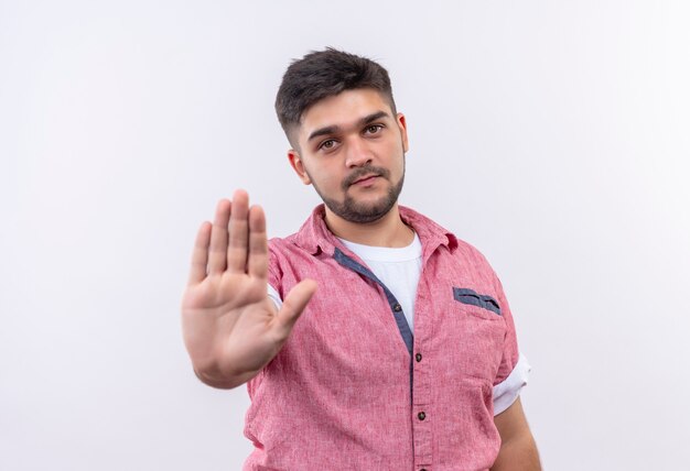 Jeune beau mec portant un polo rose faisant panneau d'arrêt avec la main debout sur un mur blanc