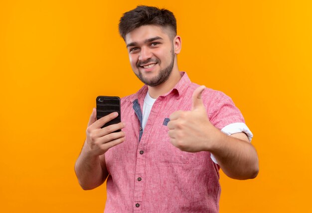 Jeune beau mec portant un polo rose faisant des pouces heureux tenant le téléphone debout sur un mur orange