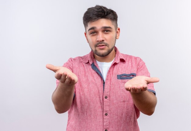 Jeune beau mec portant un polo rose indécis ne sait pas quoi faire debout sur un mur blanc