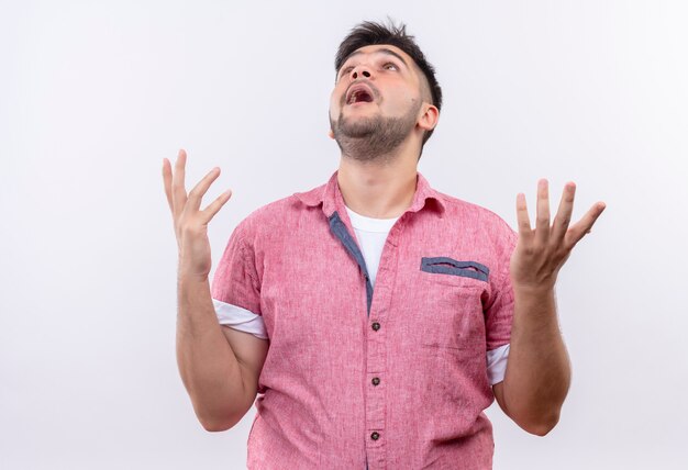 Jeune beau mec portant un polo rose llooking avec les mains levées debout sur un mur blanc