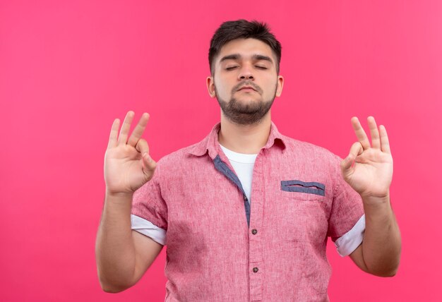 Jeune beau mec portant un polo rose méditant patiemment avec les yeux fermés debout sur le mur rose