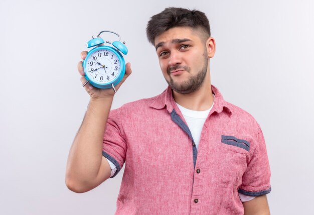 Jeune beau mec portant un polo rose montrant un réveil vous faisant face à la fin debout sur un mur blanc