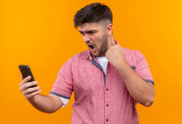 Jeune beau mec portant un polo rose à la recherche sur le téléphone faisant le poing debout sur le mur orange