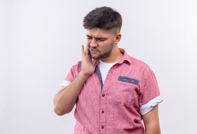 Jeune beau mec portant un polo rose souffrant de maux de dents tenant ses dents avec la main debout sur un mur blanc