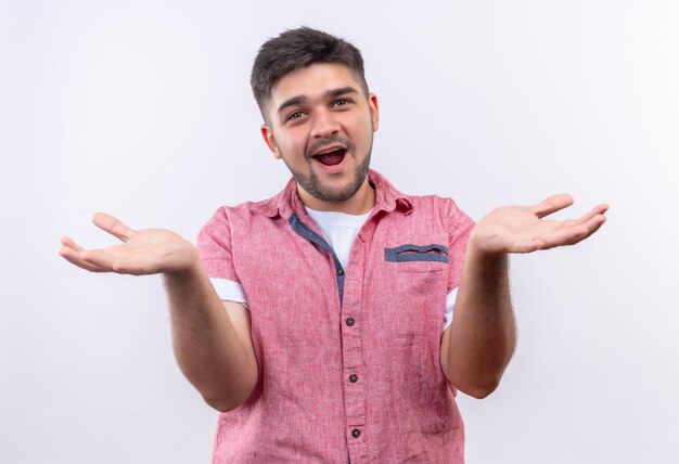 Jeune beau mec portant un polo rose souriant indécis main levée debout sur un mur blanc