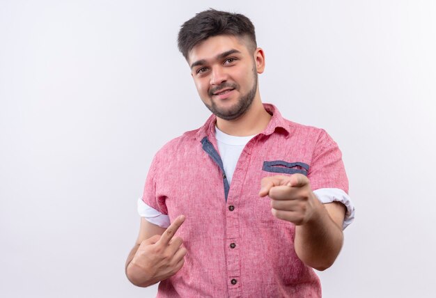 Jeune beau mec portant un polo rose souriant pointant avec l'index debout sur un mur blanc