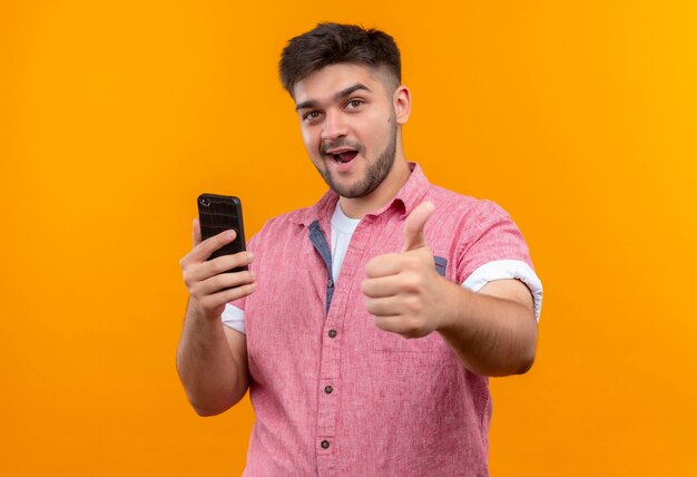 Jeune beau mec portant un polo rose tenant le téléphone faisant des pouces heureux debout sur un mur orange