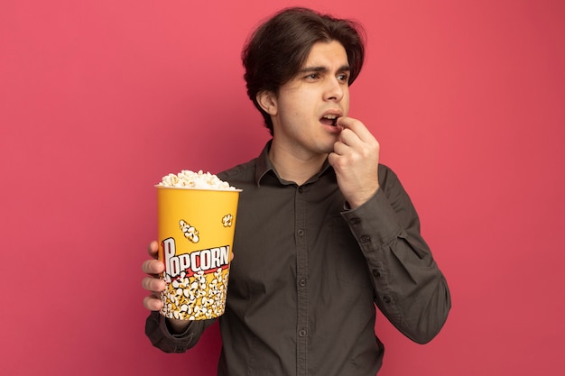 Jeune beau mec regardant côté portant un t-shirt noir tenant et essayant un seau de pop-corn isolé sur un mur rose