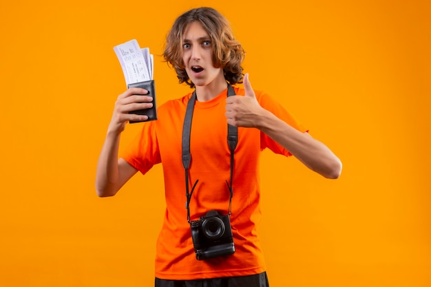 Jeune beau mec en t-shirt orange avec appareil photo tenant des billets d'avion souriant joyeusement montrant les pouces vers le haut debout sur fond jaune