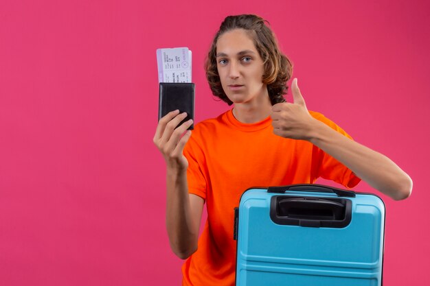 Jeune beau mec en t-shirt orange debout avec valise de voyage tenant des billets d'avion regardant la caméra positive et heureuse montrant les pouces vers le haut