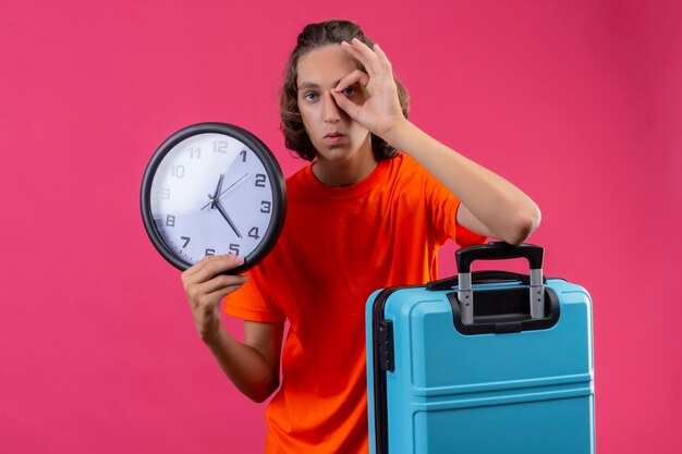 Jeune beau mec en t-shirt orange debout avec valise de voyage tenant horloge faisant signe ok à travers ce signe sur fond rose