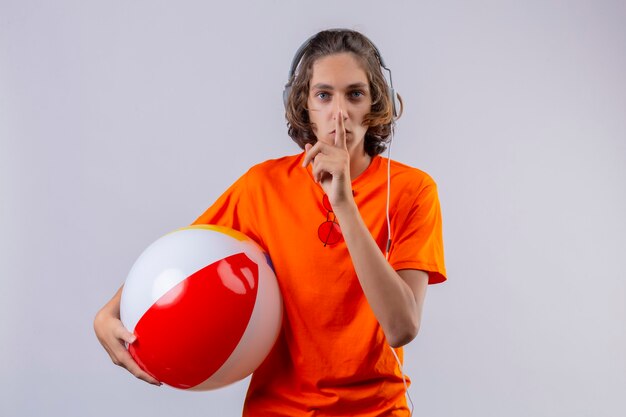 Jeune beau mec en t-shirt orange avec des écouteurs tenant ballon gonflable faisant le geste de silence avec le doigt sur les lèvres debout sur fond blanc