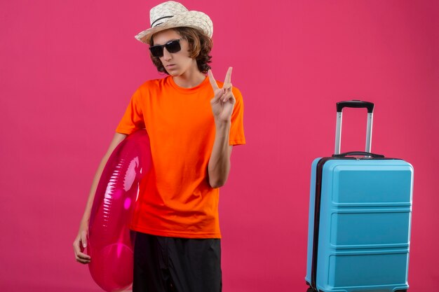 Jeune beau mec en t-shirt orange portant des lunettes de soleil noires tenant un anneau gonflable debout avec valise de voyage à la confiance montrant le signe de la victoire sur fond rose