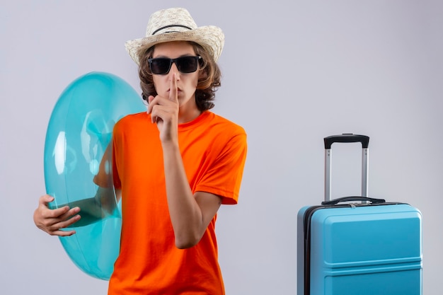 Jeune beau mec en t-shirt orange portant des lunettes de soleil noires tenant une bague gonflable faisant un geste de silence avec le doigt sur les lèvres debout avec valise de voyage sur fond blanc