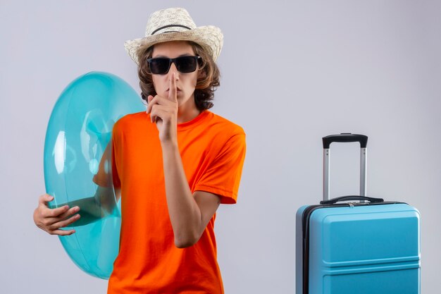 Jeune beau mec en t-shirt orange portant des lunettes de soleil noires tenant une bague gonflable faisant un geste de silence avec le doigt sur les lèvres debout avec valise de voyage