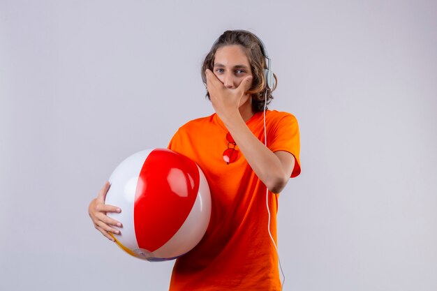 Jeune beau mec en t-shirt orange tenant ballon gonflable avec un casque étonné et surpris couvrant la bouche avec la main debout sur fond blanc