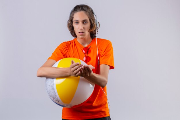 Jeune beau mec en t-shirt orange tenant ballon gonflable avec des écouteurs faisant des gestes avec les mains tenant ensemble les bras à la malheureuse debout