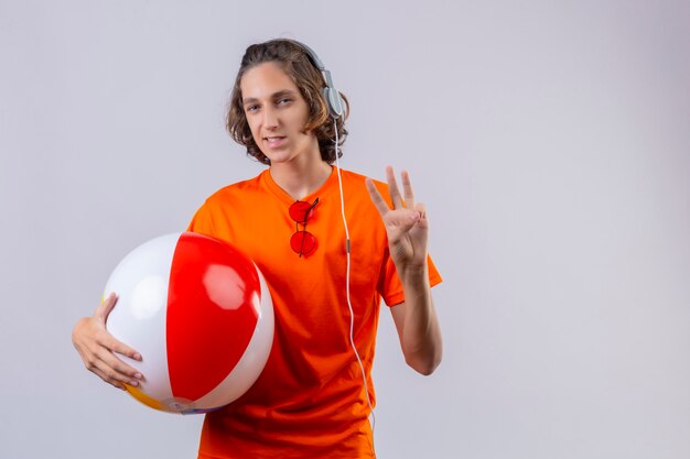 Jeune beau mec en t-shirt orange tenant ballon gonflable avec des écouteurs montrant le numéro trois souriant debout