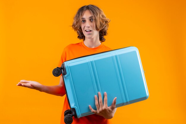 Jeune beau mec en t-shirt orange tenant une valise de voyage désemparé et confus n'ayant pas de réponse écartant les mains debout