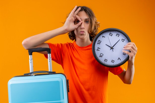 Jeune beau mec en t-shirt orange tenant valise de voyage et horloge faisant signe ok avec la main en regardant à travers ce signe avec une expression sérieuse confiante sur le visage debout sur backgroun jaune