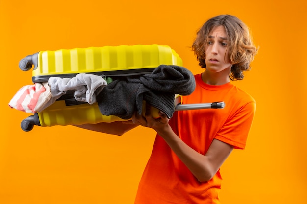 Photo gratuite jeune beau mec en t-shirt orange tenant une valise de voyage pleine de vêtements à la déçu du visage malheureux debout