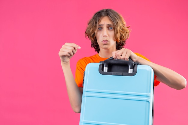 Photo gratuite jeune beau mec en t-shirt orange tenant une valise de voyage pointant avec le doigt dessus avec un visage malheureux debout