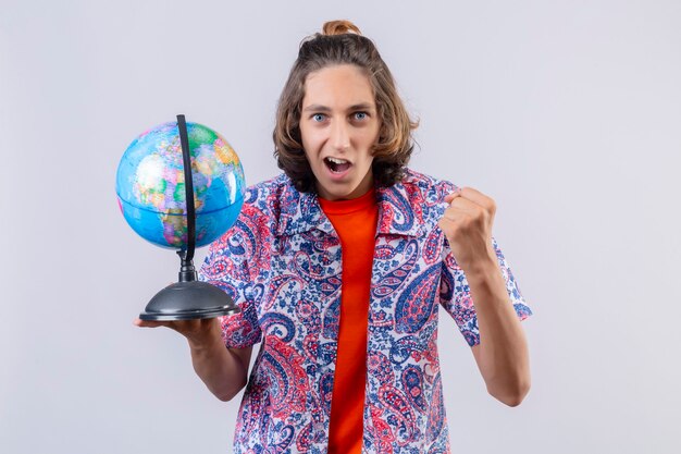 Jeune beau mec tenant un globe à la sortie et heureux de lever le poing après une victoire se réjouissant de son succès debout sur fond blanc