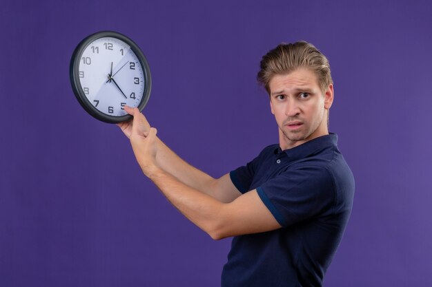 Jeune beau mec tenant horloge regardant la caméra avec une expression de confusion debout sur fond violet