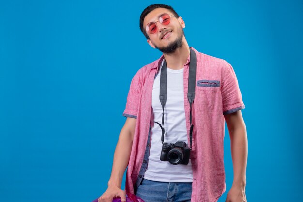 Jeune beau mec voyageur avec appareil photo portant des lunettes de soleil regardant la caméra avec un sourire confiant debout sur fond bleu