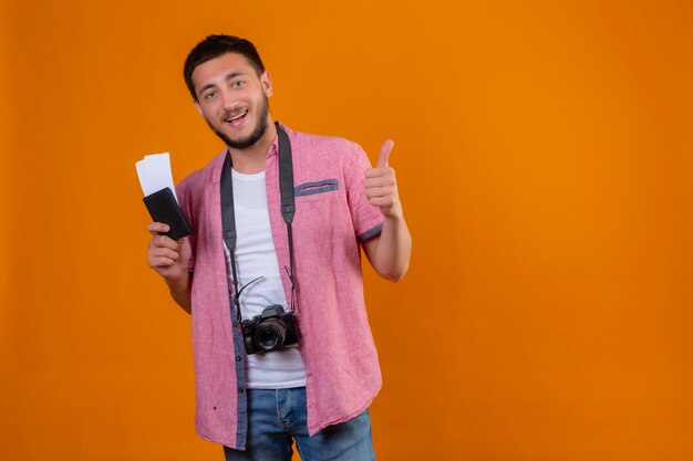 Jeune beau mec voyageur avec caméra tenant des billets d'avion regardant la caméra en souriant joyeusement montrant les pouces vers le haut debout sur fond orange