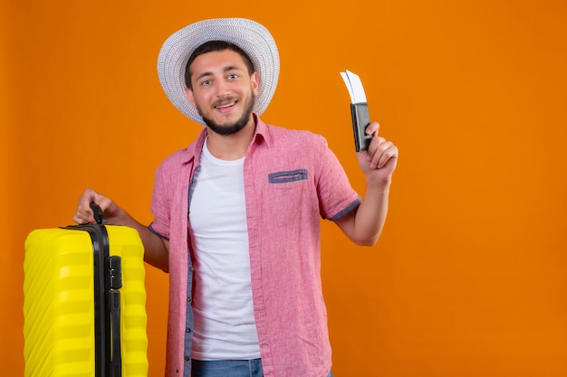 Jeune beau mec voyageur en chapeau d'été tenant valise et billets d'avion à la confiance en soi souriant joyeusement prêt à voyager debout sur fond orange