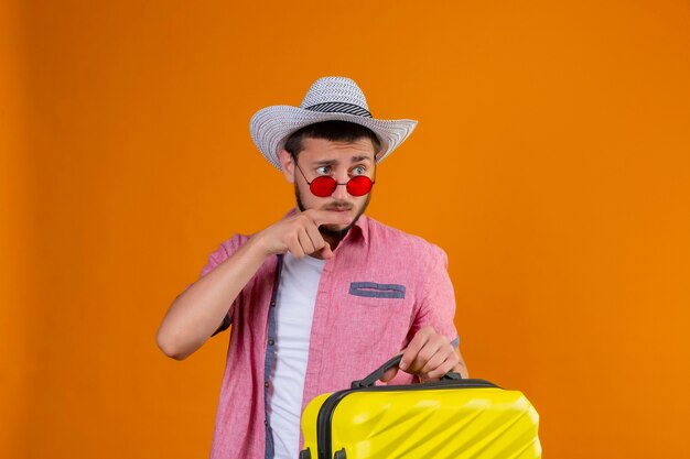 Jeune beau mec voyageur portant des lunettes de soleil en chapeau d'été tenant valise à côté en pointant avec le doigt vers quelque chose avec une expression suspecte debout sur fond orange