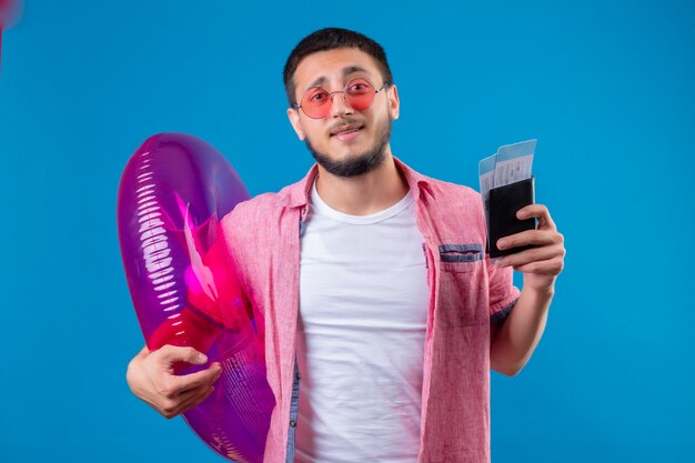 Jeune beau mec voyageur portant des lunettes de soleil tenant un anneau gonflable et des billets d'avion à la confusion debout sur fond bleu