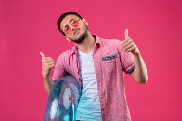 Jeune beau mec voyageur portant des lunettes de soleil tenant un anneau gonflable à la joyeuse et heureuse sourire joyeusement montrant les pouces vers le haut debout sur fond rose