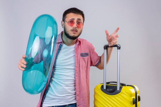 Jeune beau mec voyageur portant des lunettes de soleil tenant un anneau gonflable regardant la caméra avec une expression de confusion sur le visage debout avec valise de voyage sur fond blanc