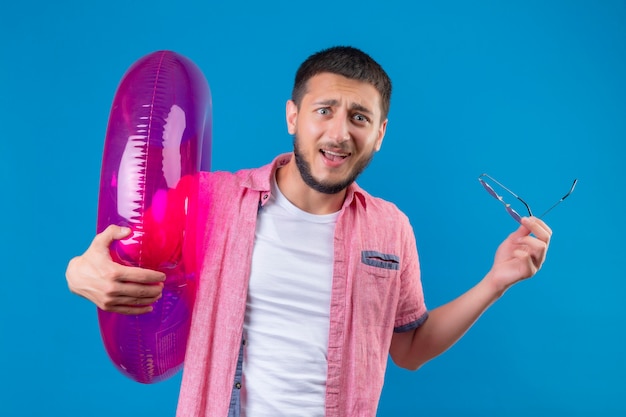 Jeune beau mec voyageur tenant un anneau gonflable regardant la caméra avec une expression de confusion sur le visage debout sur fond bleu